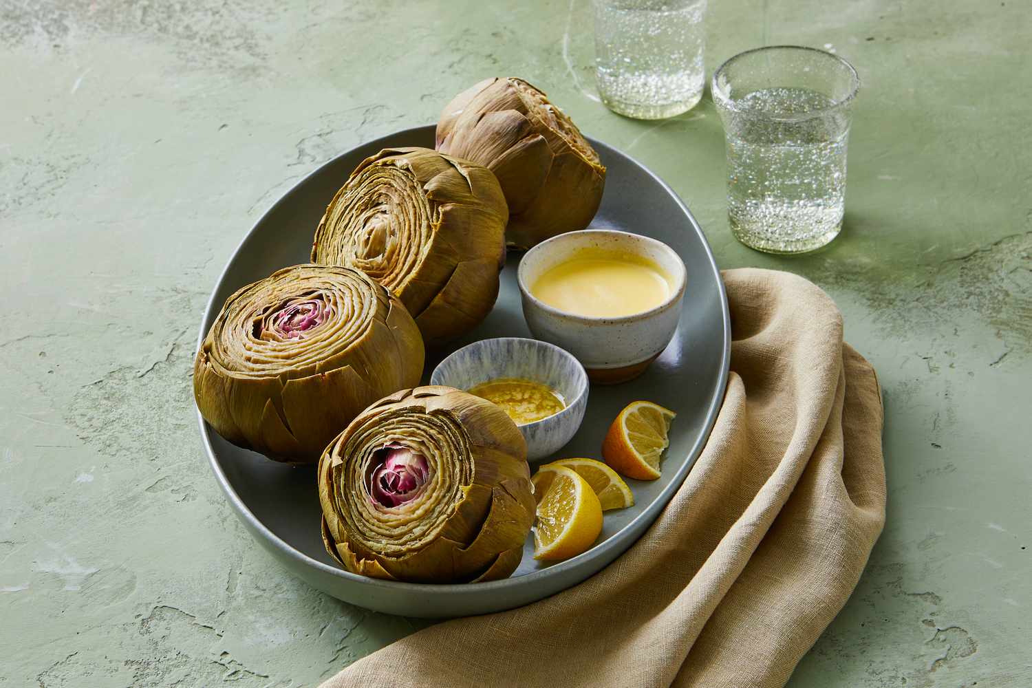 A steamed whole artichoke on a small plate with a bowl of lemon-yogurt dip and a lemon wedge